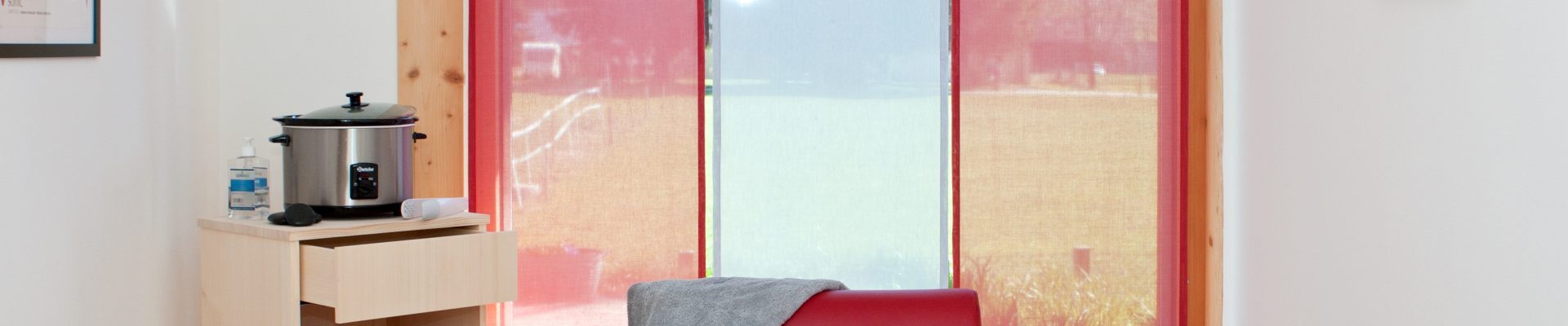 Kräuterkocher neben roter Therapieliege vor farbigen Fensterrollos